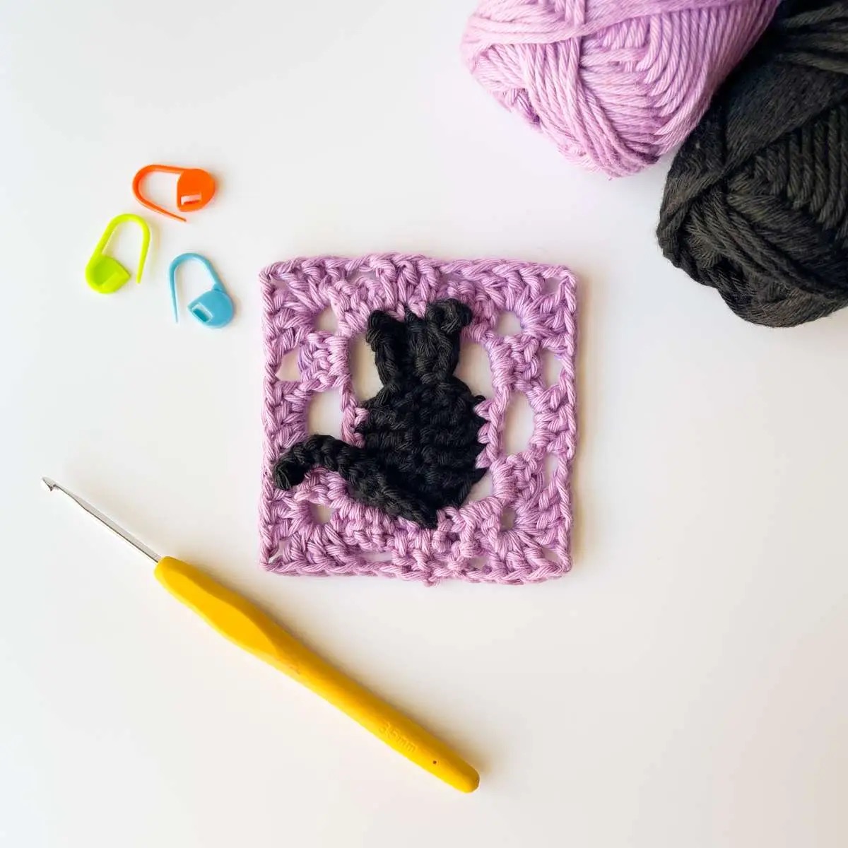 Pink crochet granny square with a black cat motif, hook and yarn