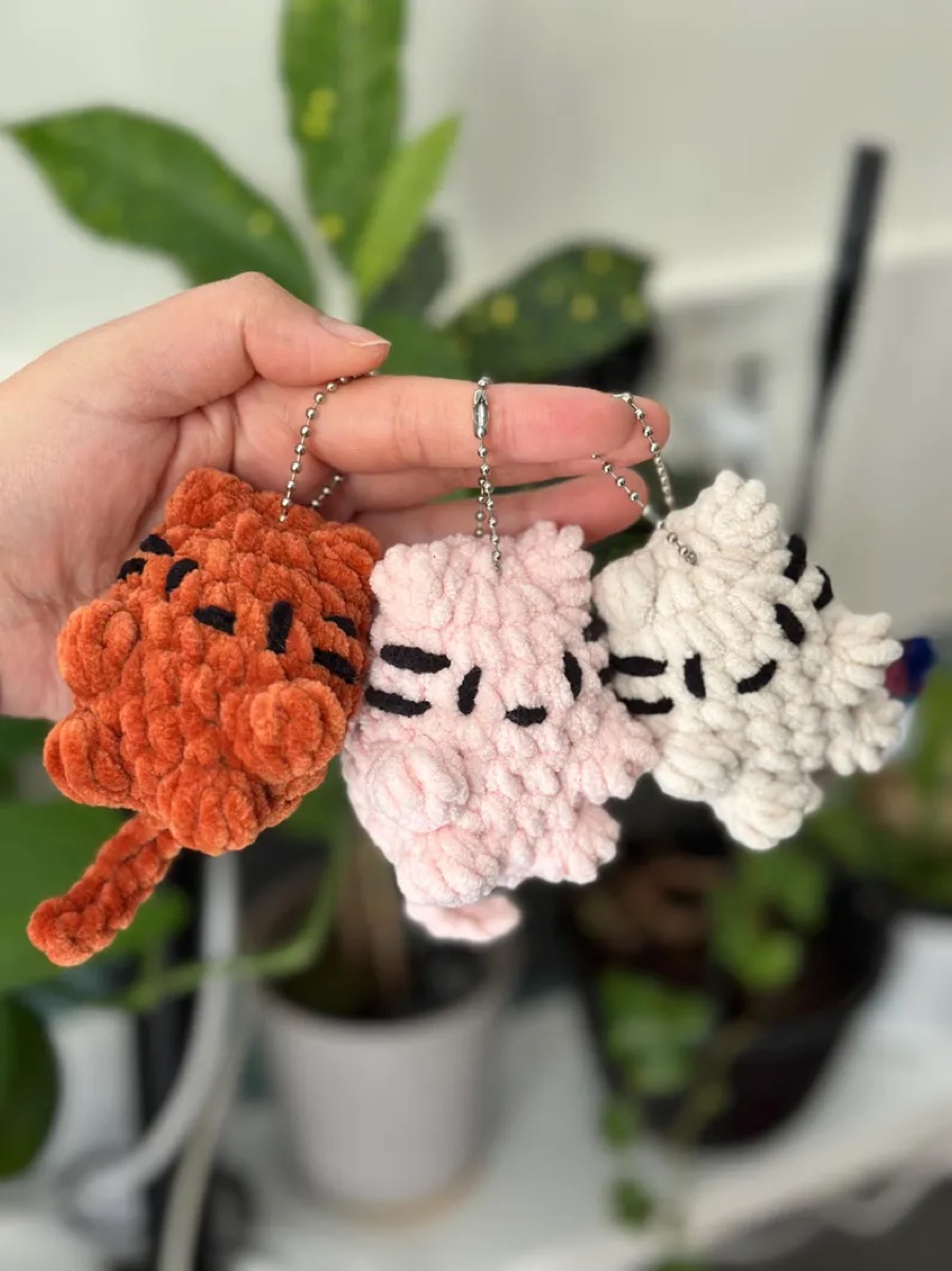 Three chunky crochet cat keychains in orange, pink and cream held in a hand
